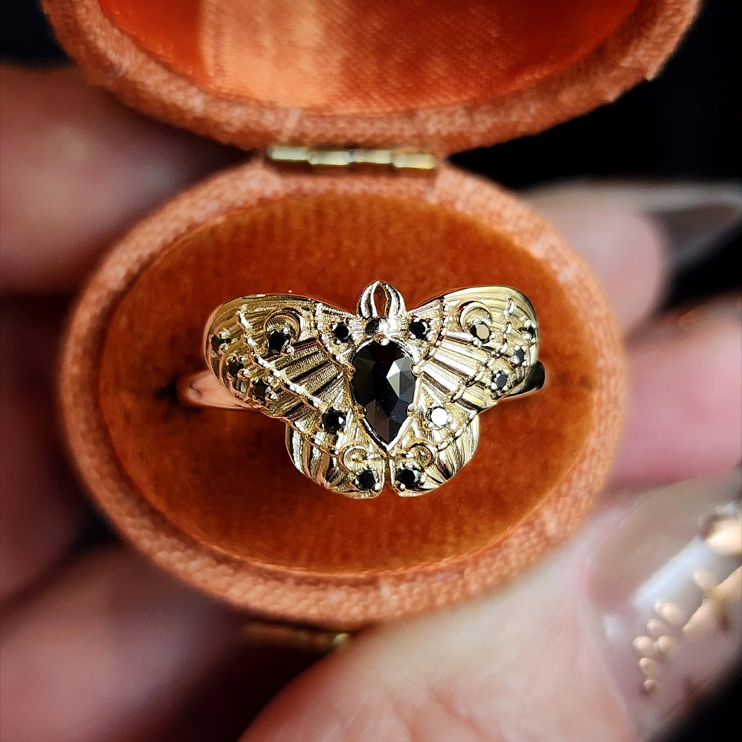 Gold butterfly ring with black gemstone in a jewelry box held by a hand.