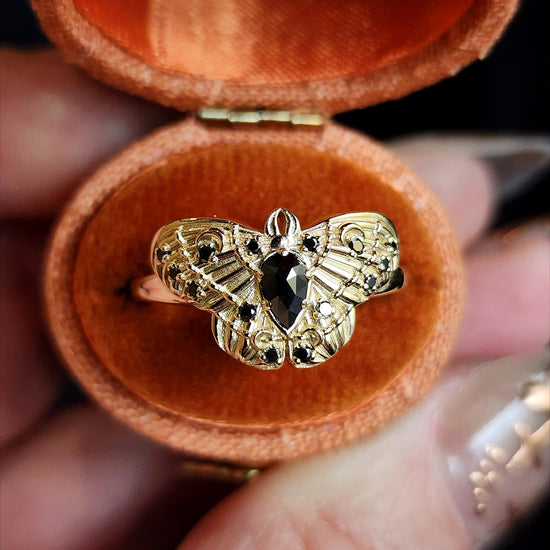 Gold butterfly ring with black gemstone in a jewelry box held by a hand.