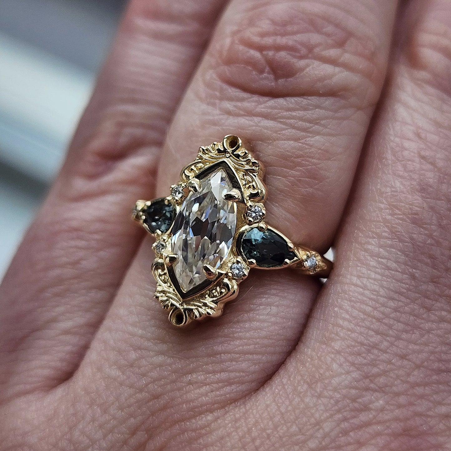 This Odette ring is a stunning piece, centered around a striking 1.20ct Moissanite Marquise, featuring an Old Mine Cut This center stone is beautifully complemented by Natural Australian Pear Sapphires, totaling .43ct, and accented with .05ct VS natural white diamonds Crafted in 14kt yellow gold image 10