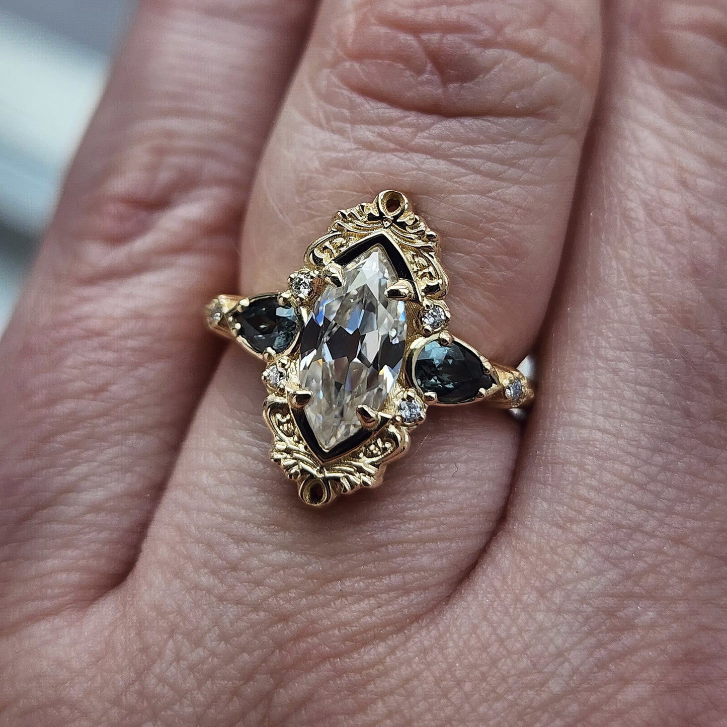 This Odette ring is a stunning piece, centered around a striking 1.20ct Moissanite Marquise, featuring an Old Mine Cut This center stone is beautifully complemented by Natural Australian Pear Sapphires, totaling .43ct, and accented with .05ct VS natural white diamonds Crafted in 14kt yellow gold image 9