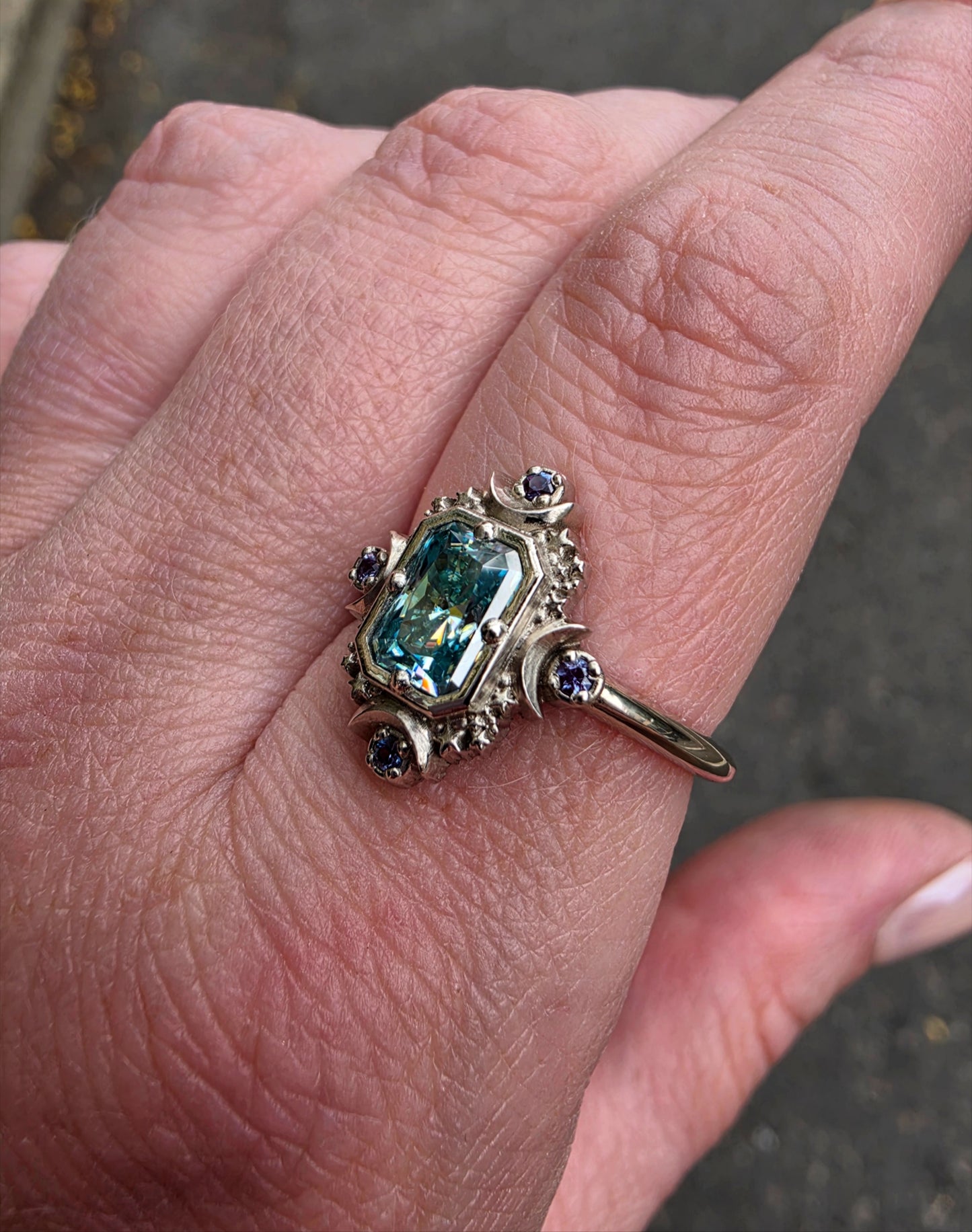 A close-up of a hand wearing a vintage-style ring with a rectangular light blue gemstone and small dark blue accent stones on a silver-toned band.