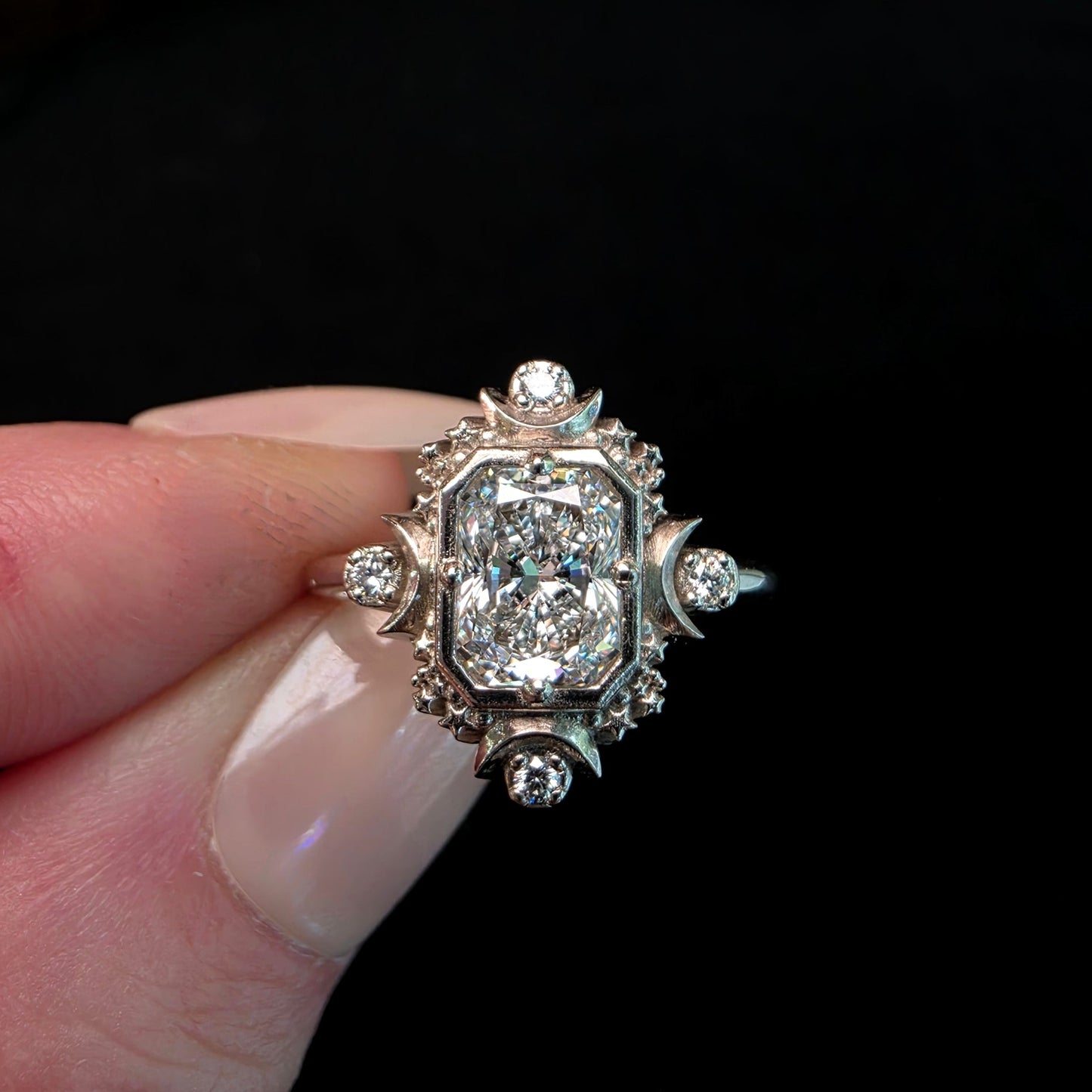 An antique-style diamond ring with a large rectangular cushion-cut center stone and smaller diamonds, held between fingers against a dark background.