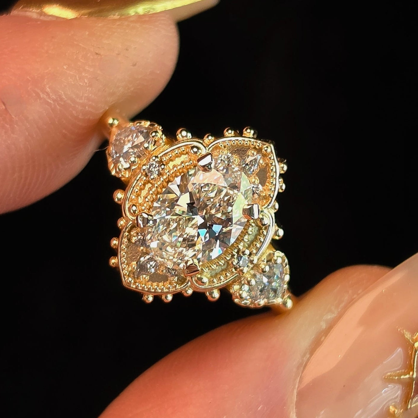 A close-up of a vintage-style gold ring with a large clear central gemstone and smaller surrounding stones, held between two fingers against a black background.