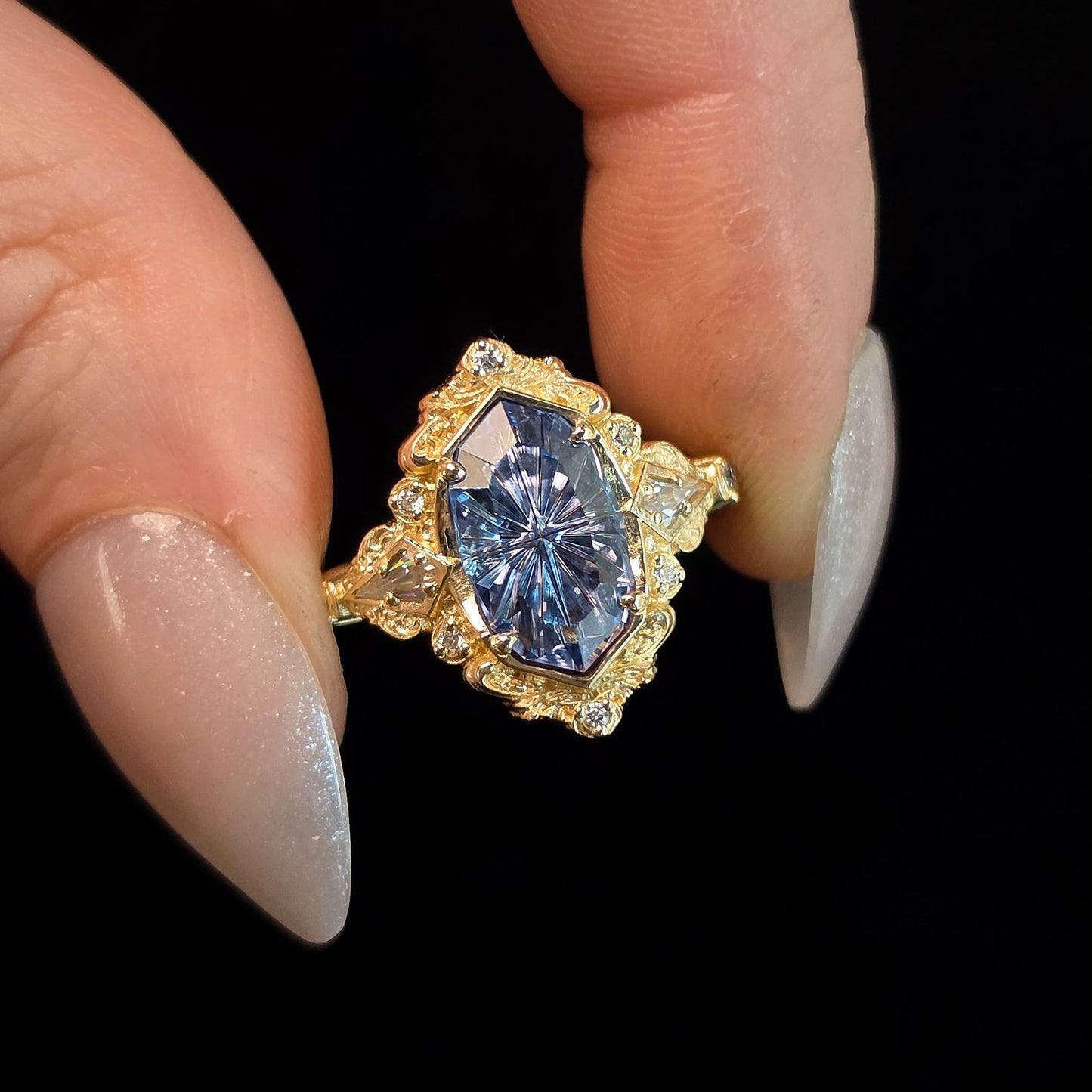 A close-up of a hand with long nude manicured nails holding a yellow gold ring featuring a large oval-cut blue gemstone surrounded by small clear accent stones, set against a solid black background.