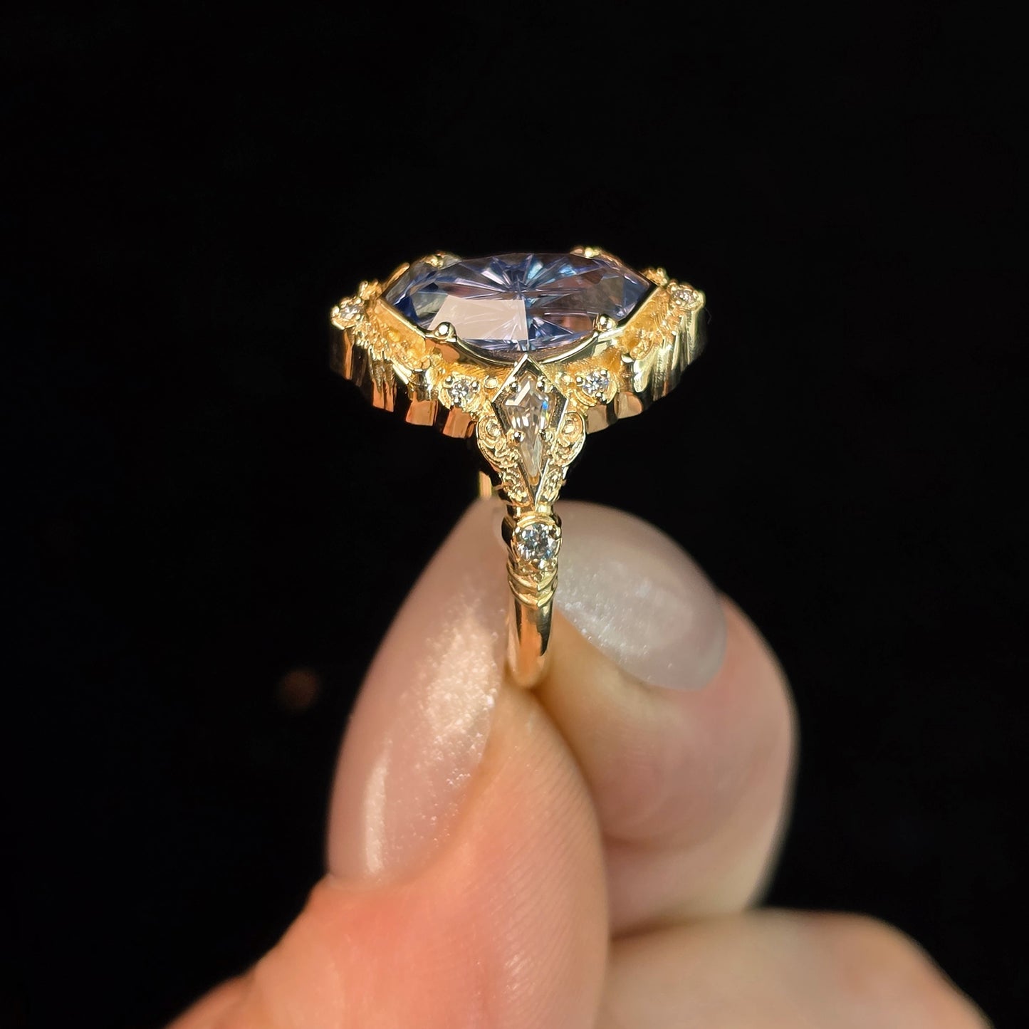 A close-up of a hand holding a vintage-style gold ring with a marquise-cut blue gemstone and small accent diamonds, set against a solid black background.