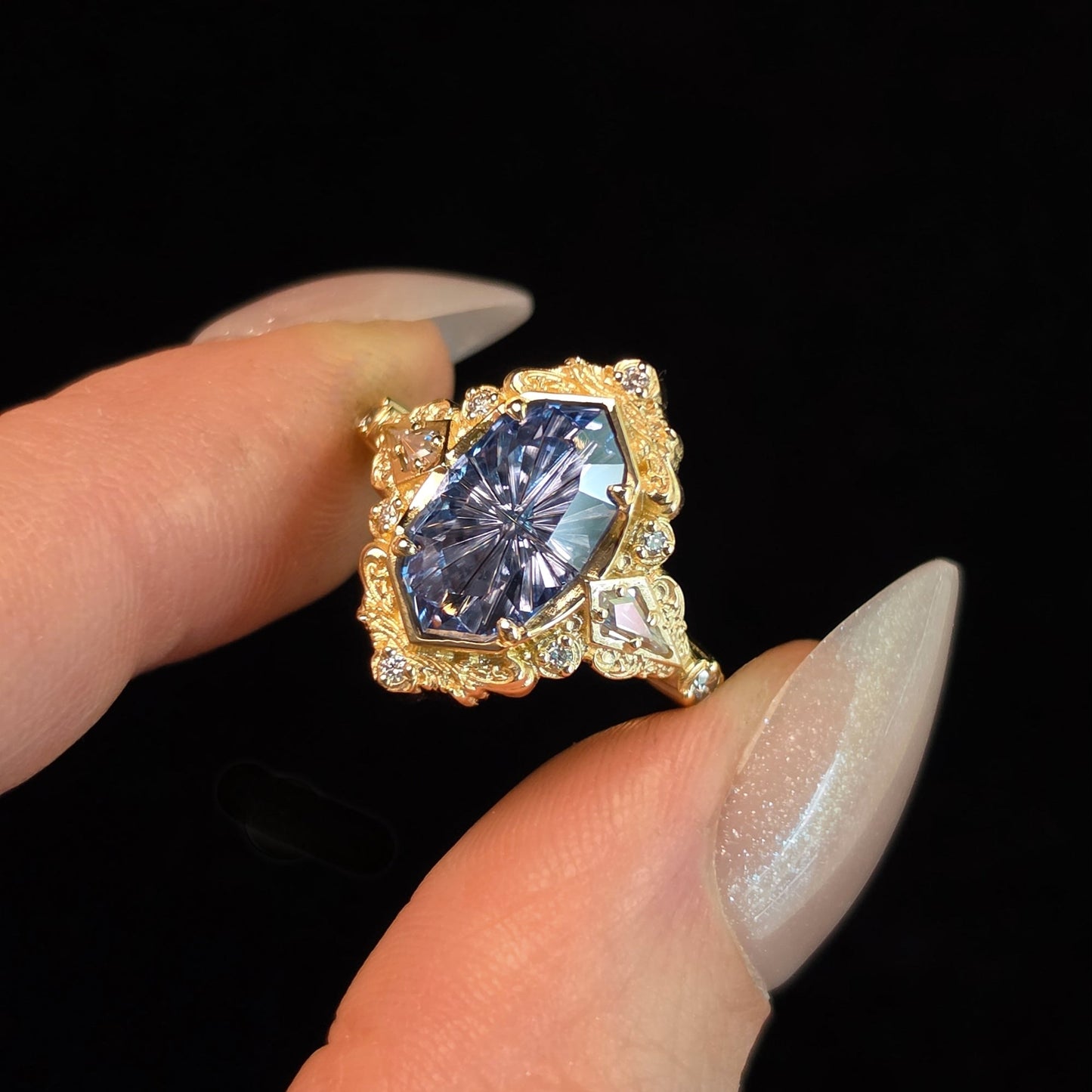 A close-up of a hand with long, manicured nails holding a gold ring featuring a marquise-cut blue gemstone surrounded by small clear stones, set against a solid black background.