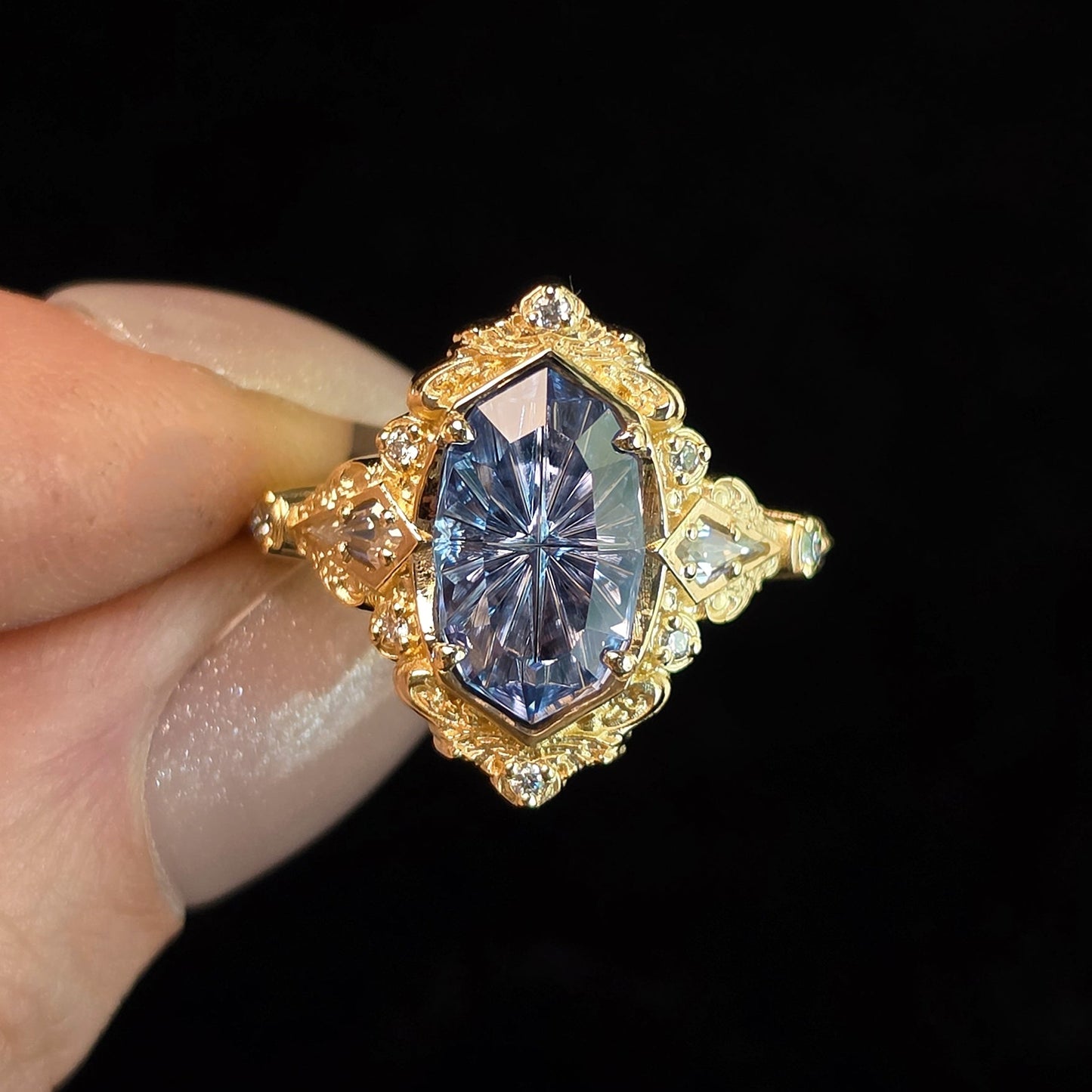 A gold ring with a large marquise-cut blue gemstone and small clear accent stones, held between fingers against a solid black background.