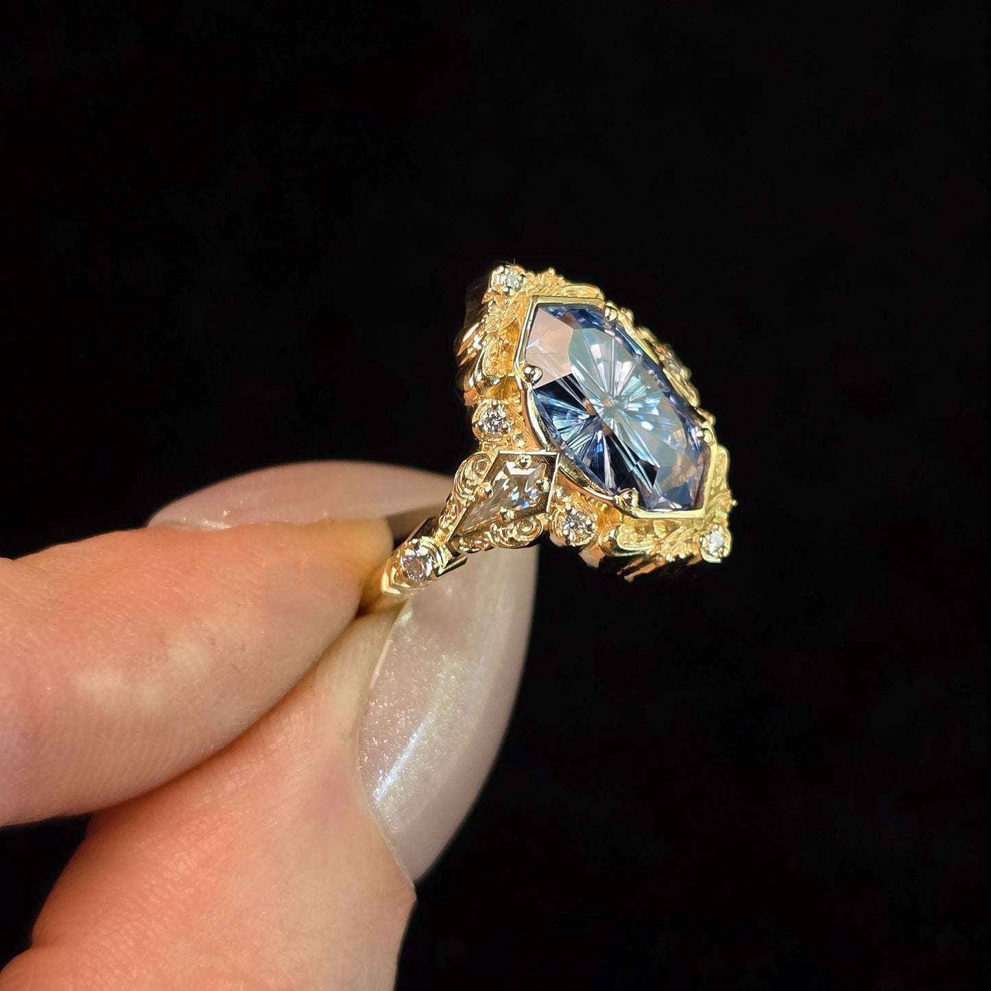 A gold ring with a large marquise-cut blue gemstone and small clear accent stones, held between fingers against a solid black background.