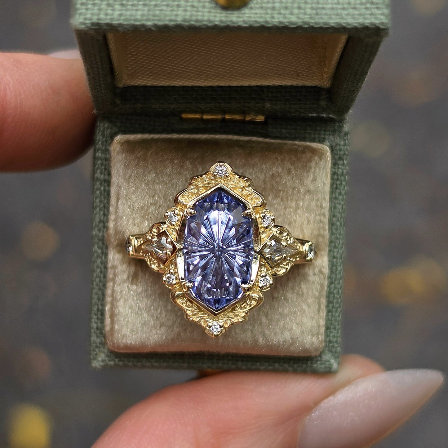 A yellow gold ring with a large oval blue gemstone and small clear accent stones, displayed in an open sage green jewelry box held by a hand.