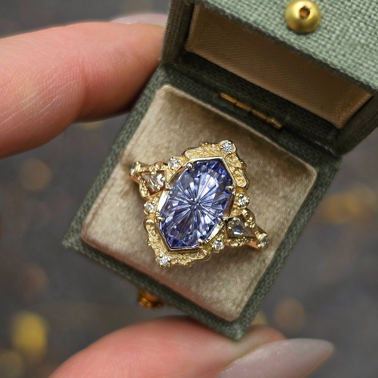 A close-up of a hand holding an open green jewelry box containing a gold ring with a large marquise-cut blue gemstone and small clear accent stones, against a blurred outdoor background.