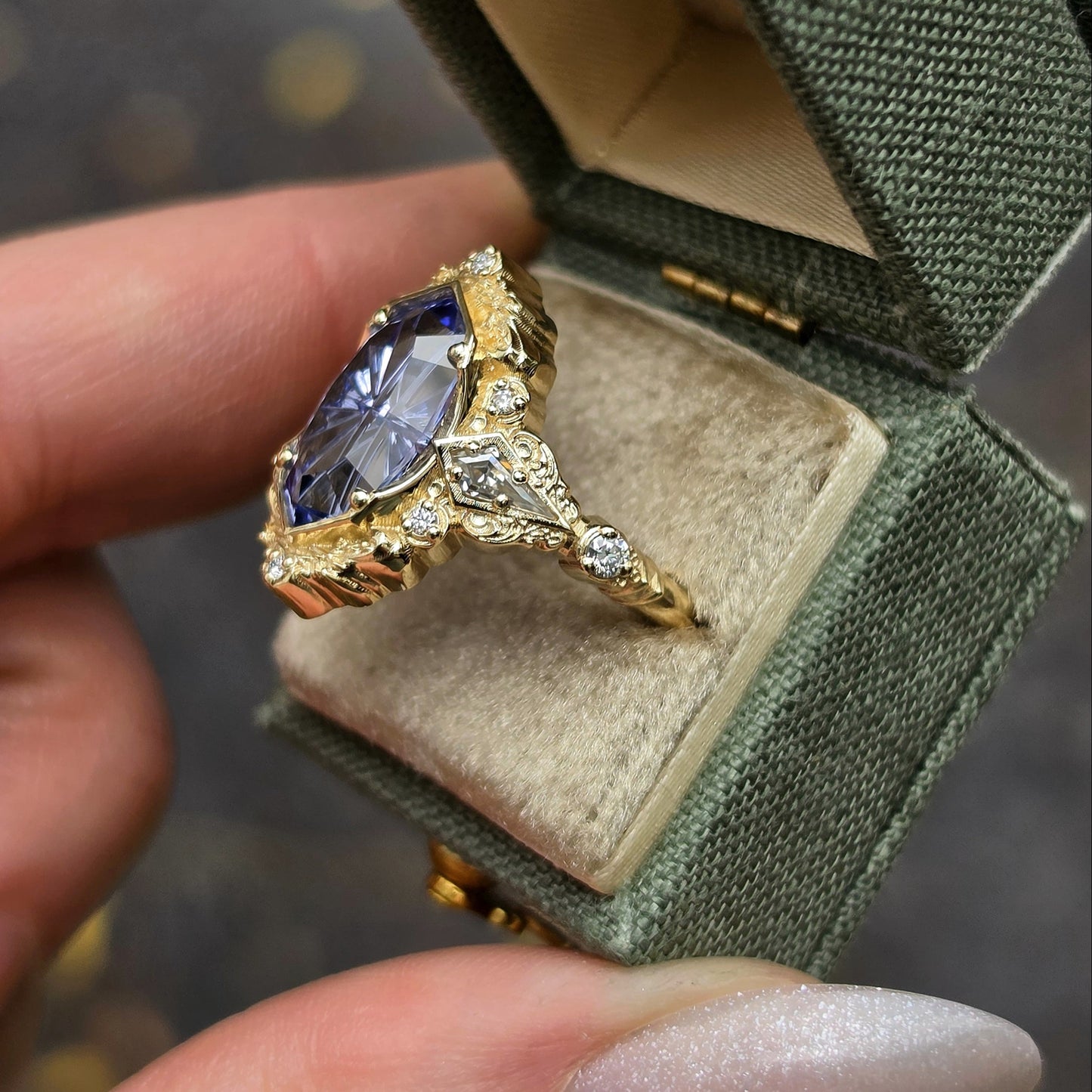A gold ring with a large oval blue gemstone and small clear accent stones, displayed in an open green jewelry box held by a hand.
