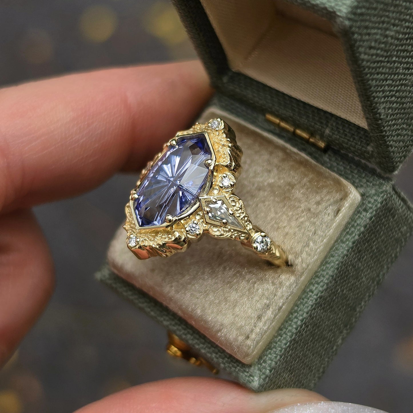 A close-up of a gold ring with a large marquise-cut blue gemstone and small clear accent stones, displayed inside an open textured green ring box held by a hand.