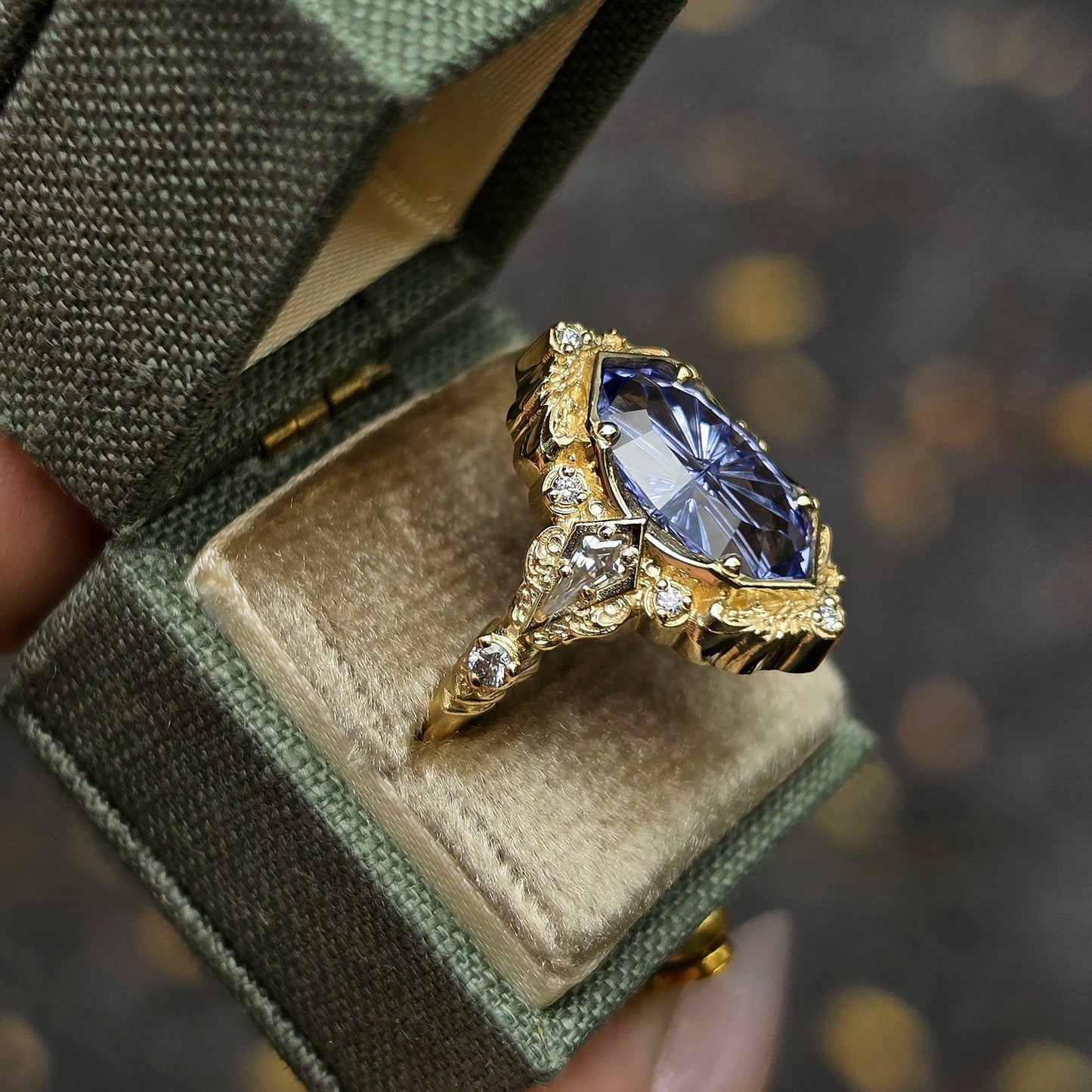 A gold ring with a large oval blue gemstone and small clear accent stones, displayed inside an open textured green ring box held by a hand.