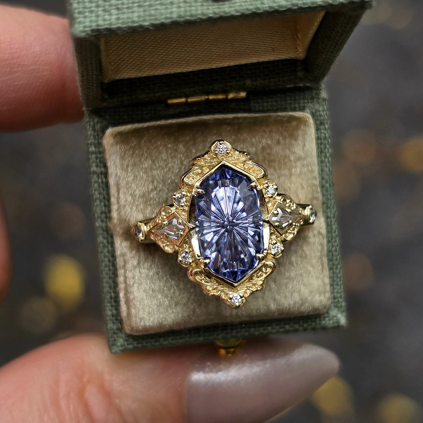 A vintage-style gold ring with a marquise-cut blue gemstone and small clear accent stones, displayed inside an open green fabric ring box held by a hand.