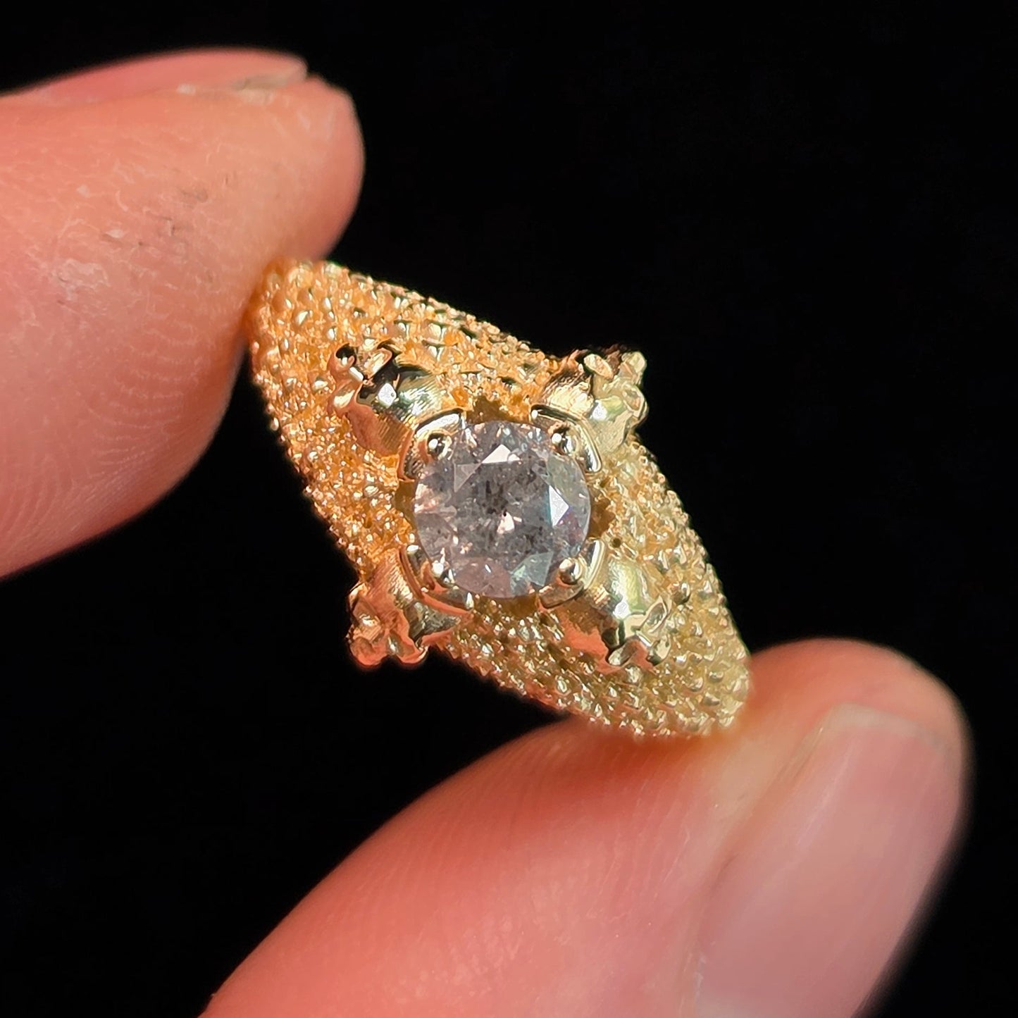 A gold ring with a large clear center stone and textured pavé detailing, held between two fingers against a solid black background.