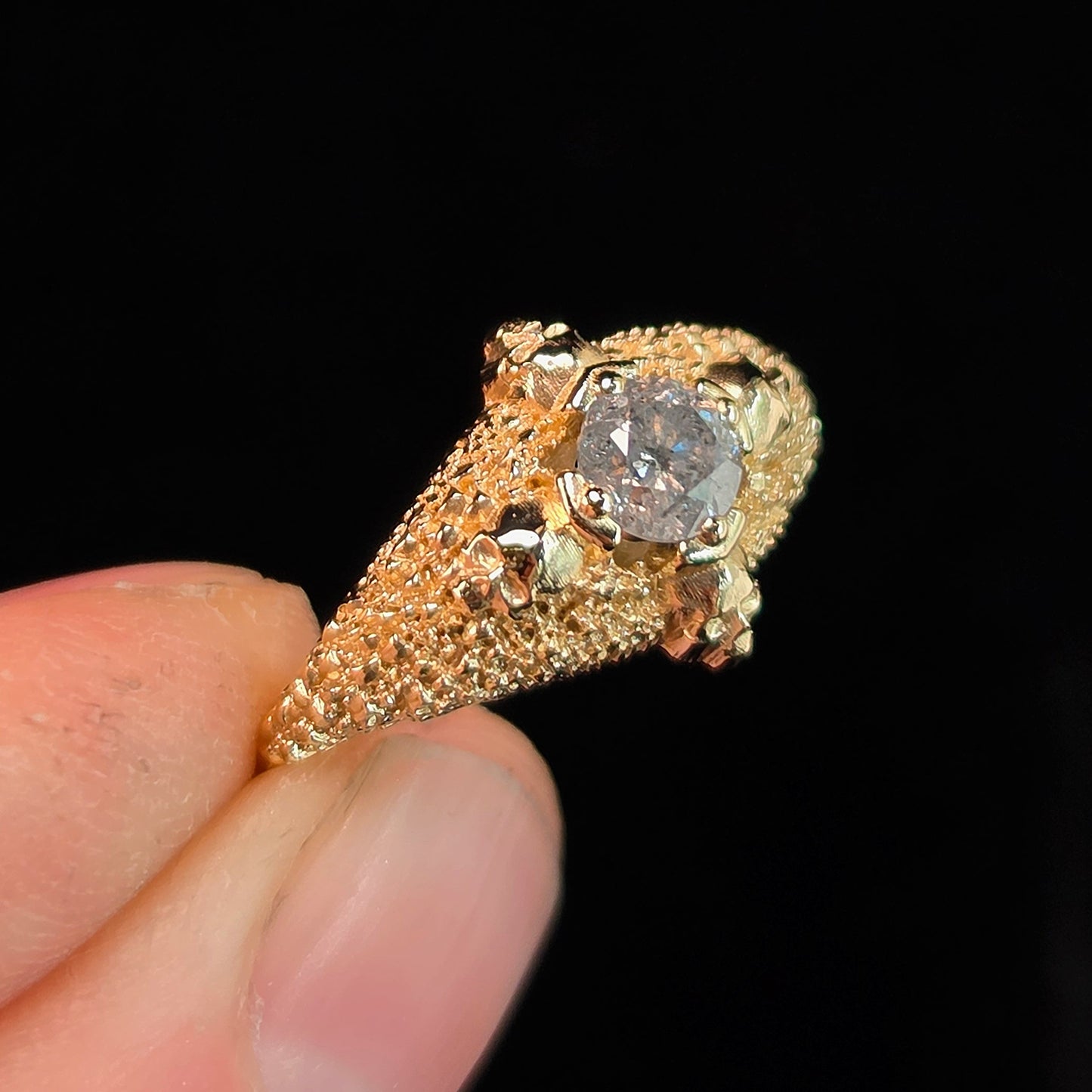 A close-up of a hand holding a yellow gold ring with a textured pavé band and a central clear gemstone, set against a solid black background.