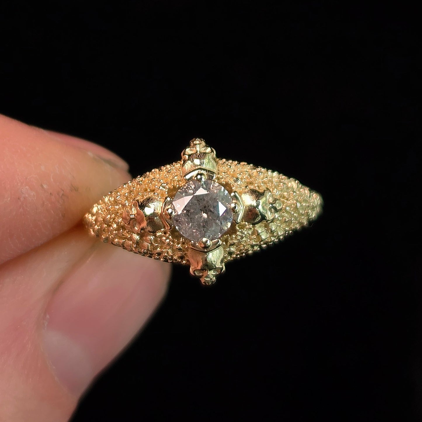 A vintage-style diamond ring with a textured gold band and a central round diamond, held between fingers against a solid black background.