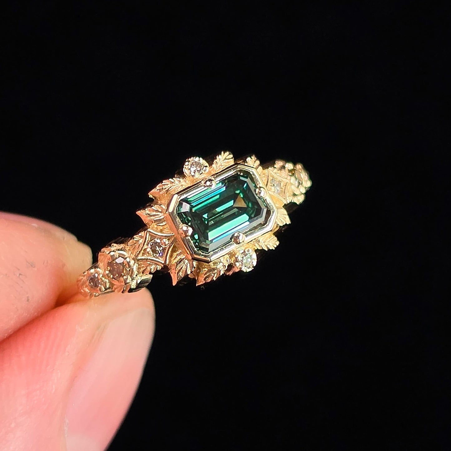 A gold ring with a rectangular emerald-cut green gemstone and small clear accent stones, held between fingers against a solid black background.