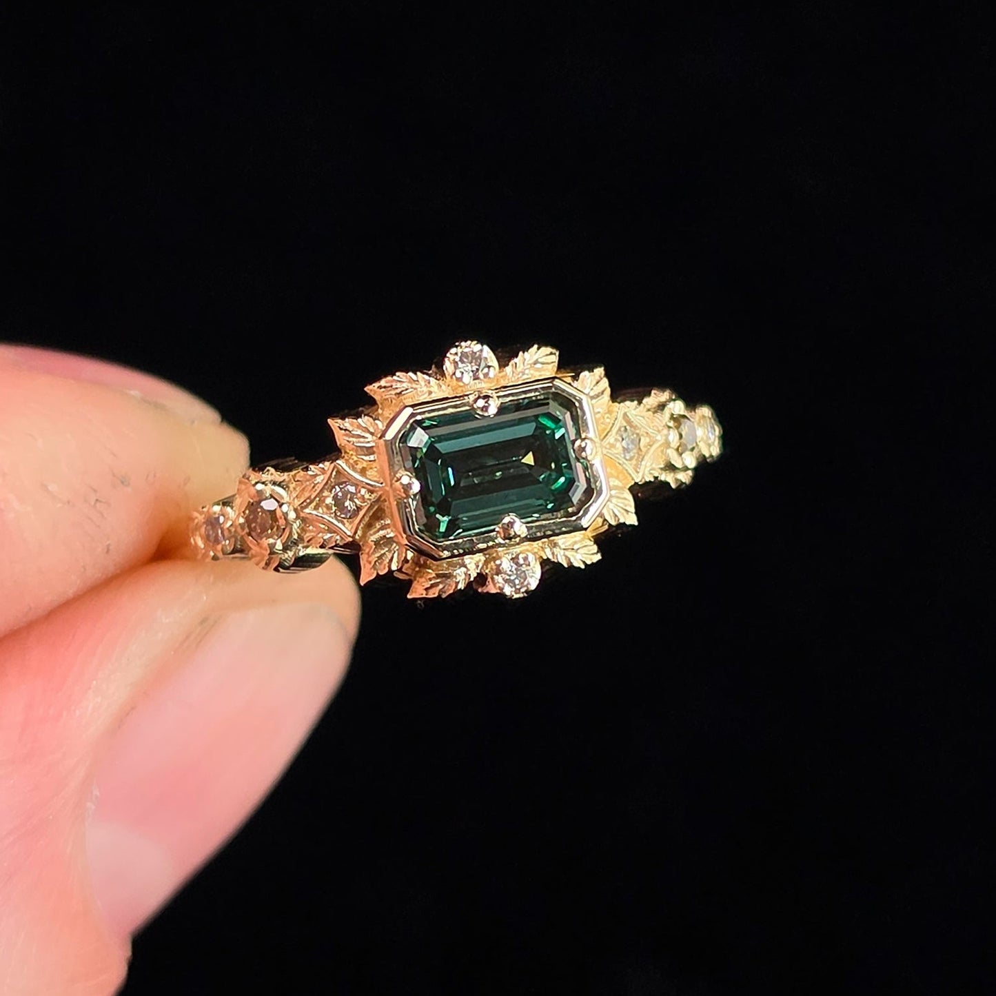 A gold ring with a rectangular green emerald-cut center stone and small clear accent stones, held between fingers against a solid black background.