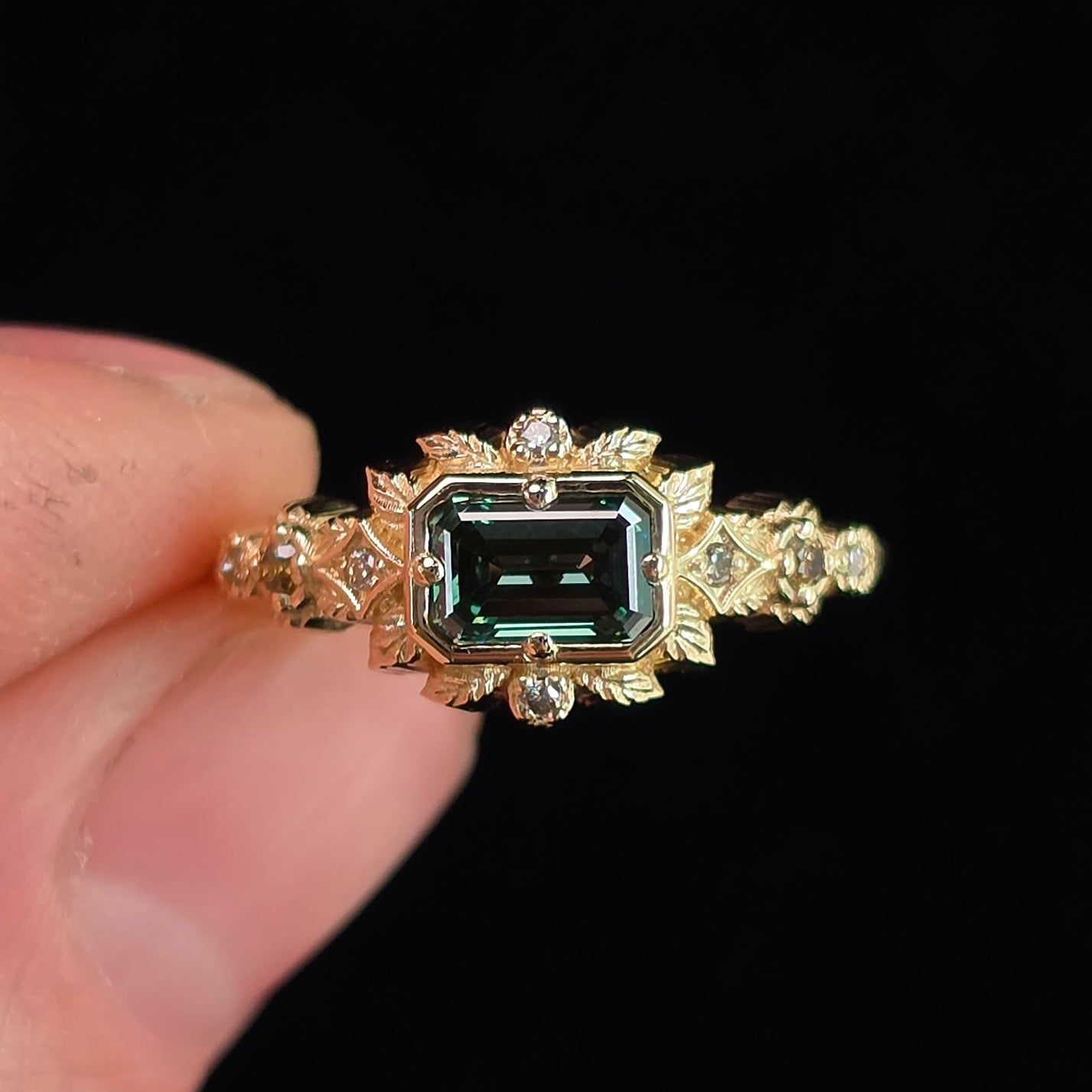 A gold ring with a rectangular emerald-cut green gemstone and small clear accent stones, held between fingers against a solid black background.