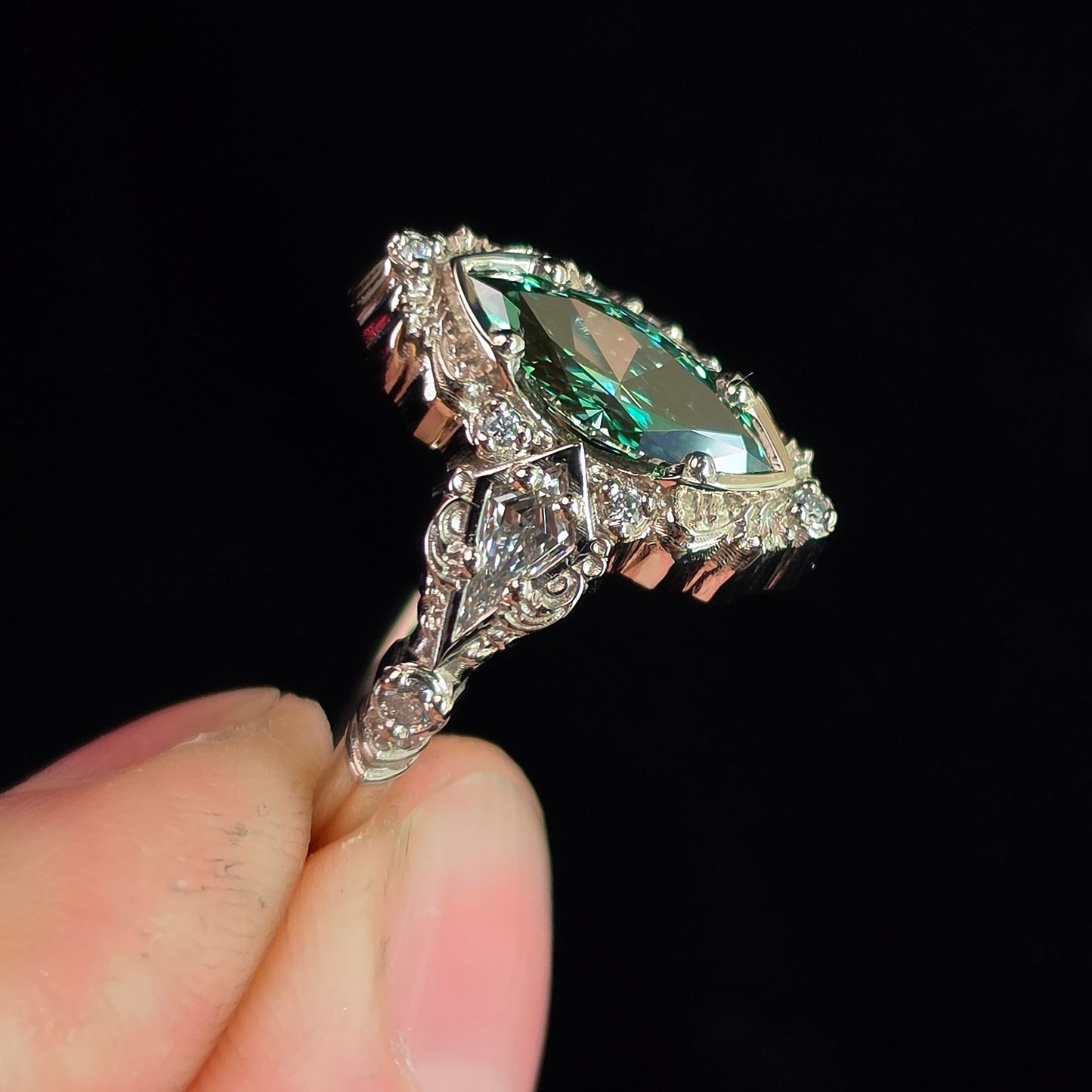 A close-up of a hand holding a silver ring with a marquise-cut green gemstone and small clear accent stones, set against a solid black background.