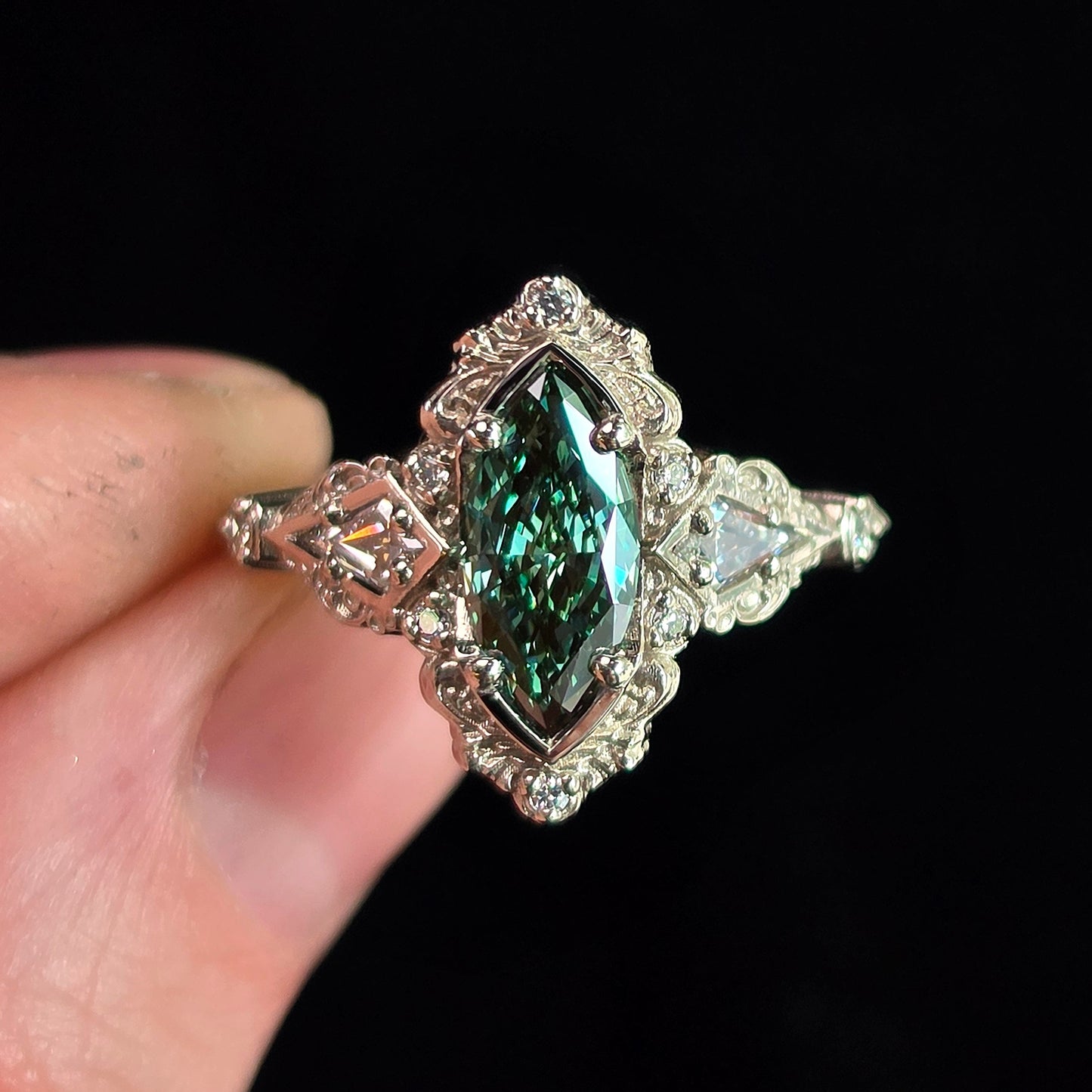 A silver-toned ring with a marquise-cut green gemstone and clear accent stones, held between fingers against a solid black background.