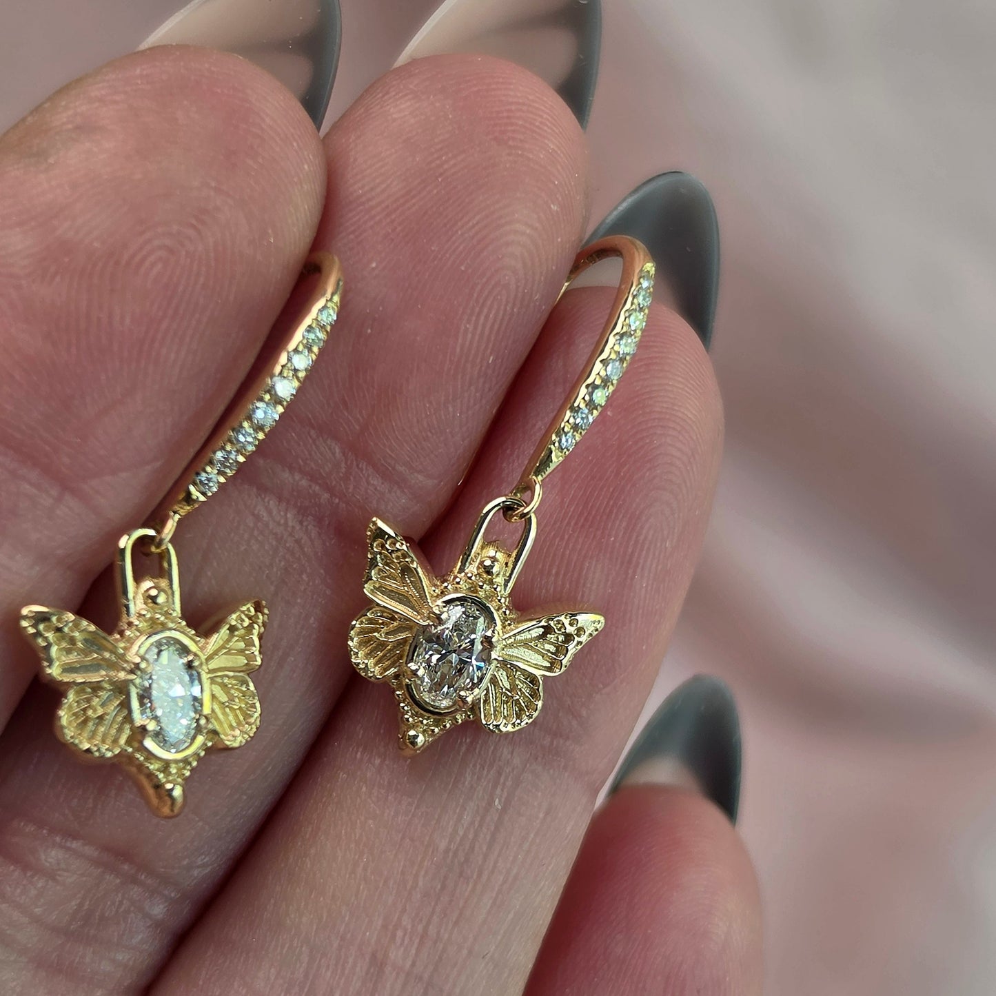 A close-up of a hand holding a pair of gold hoop earrings with butterfly charms and small clear stones, against a soft pink background.