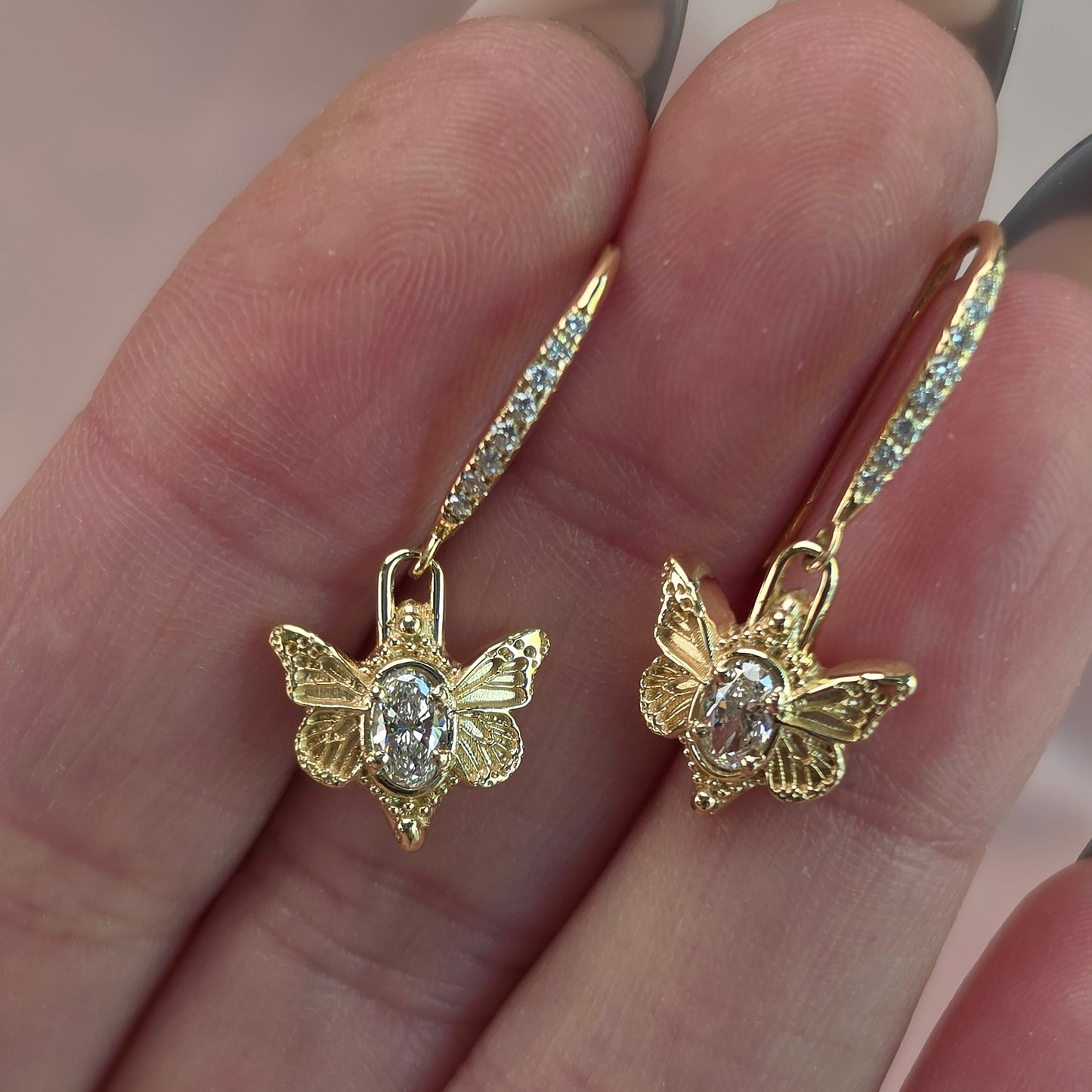 A pair of gold dangle earrings with butterfly-shaped pendants and small clear stones, shown resting on a person's fingers.