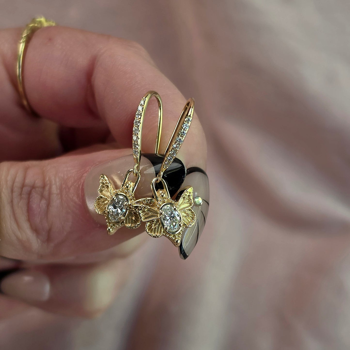 A close-up of a hand holding a pair of gold butterfly hoop earrings with clear gemstones, against a soft pink fabric background.