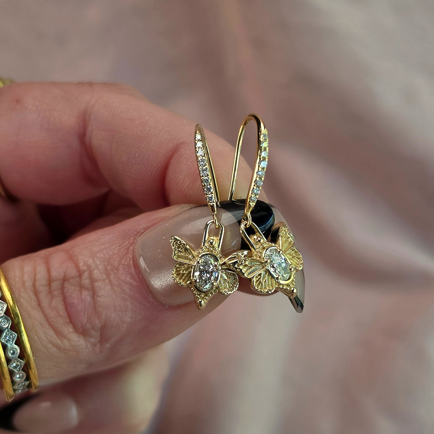 A close-up of a hand holding a pair of gold butterfly hoop earrings with clear gemstones, against a soft pink fabric background.