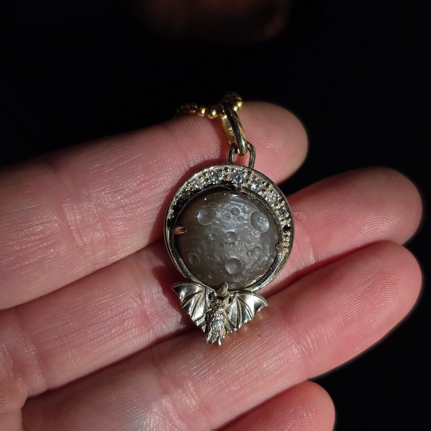 A close-up of a hand holding a silver pendant with a gray stone and a gold chain against a dark background.