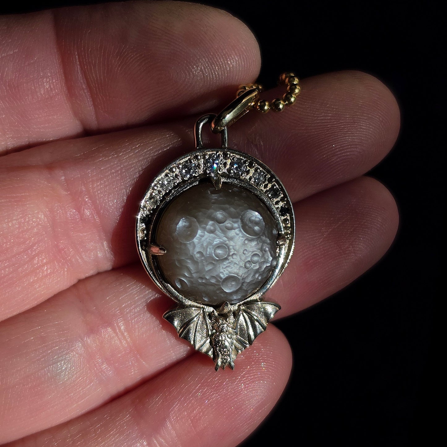 A close-up of a hand holding a silver-toned pendant with a crescent moon design and a small gold-toned clasp, set against a dark background.