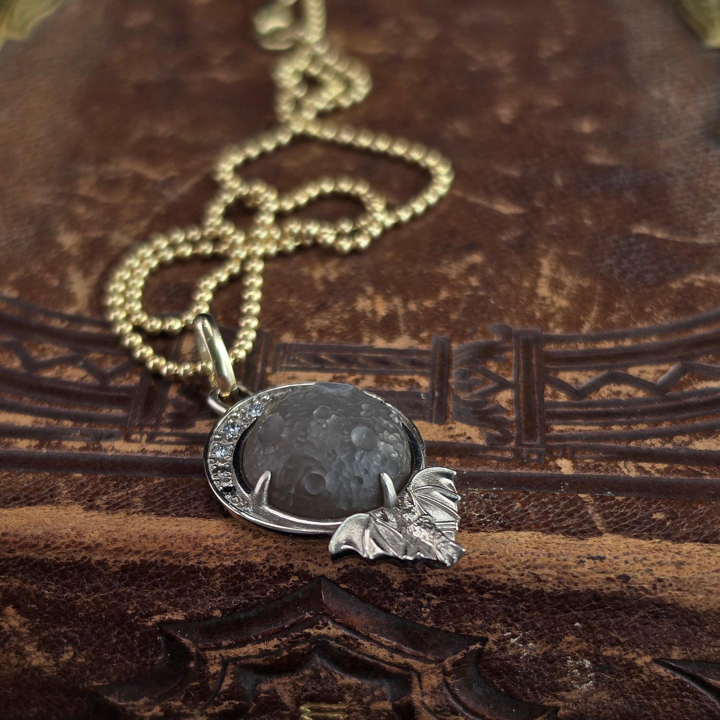 A close-up of a necklace with a round dark gray stone pendant and a silver-toned leaf charm, resting on a textured brown leather surface.