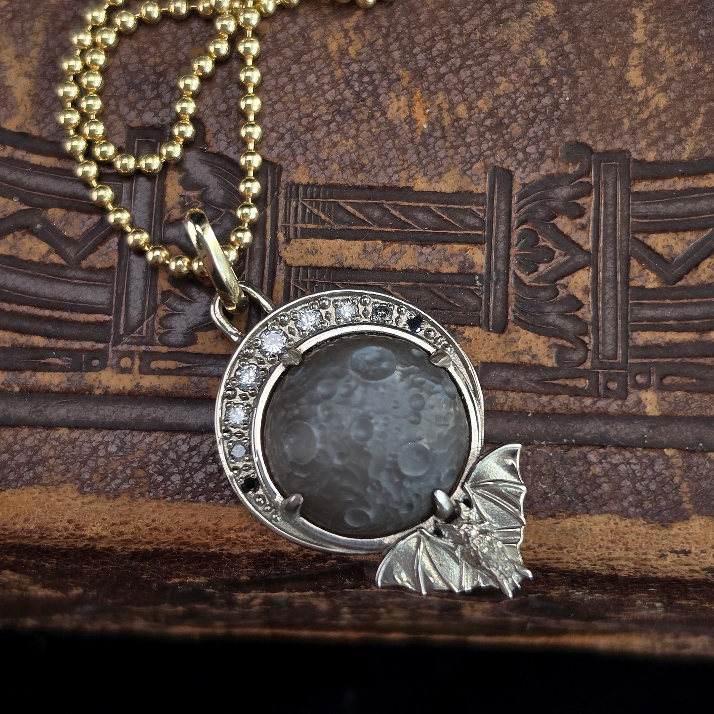 A gothic-style pendant necklace featuring a silver crescent moon with clear rhinestones and a small silver bat charm, set against a dark gray stone insert, hanging from a gold ball chain on a textured brown leather background.