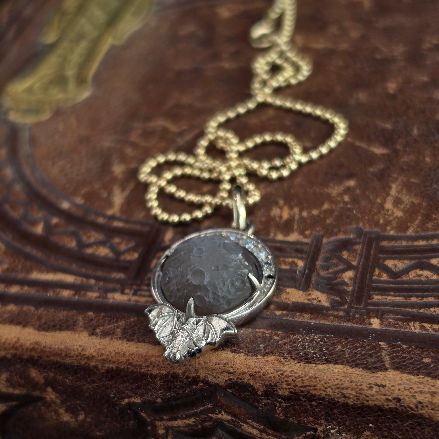 A gothic-style pendant necklace featuring a round dark gray stone and a silver bat charm, resting on an antique brown leather book.