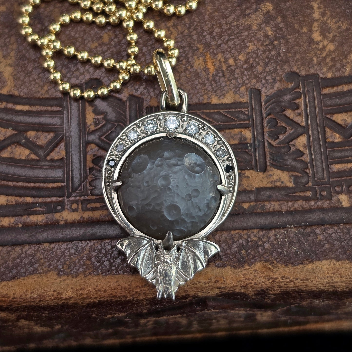 A silver-toned pendant necklace featuring a moon and bat design, with a textured dark gray stone in the center and small clear stones around the top, displayed on a brown textured background.