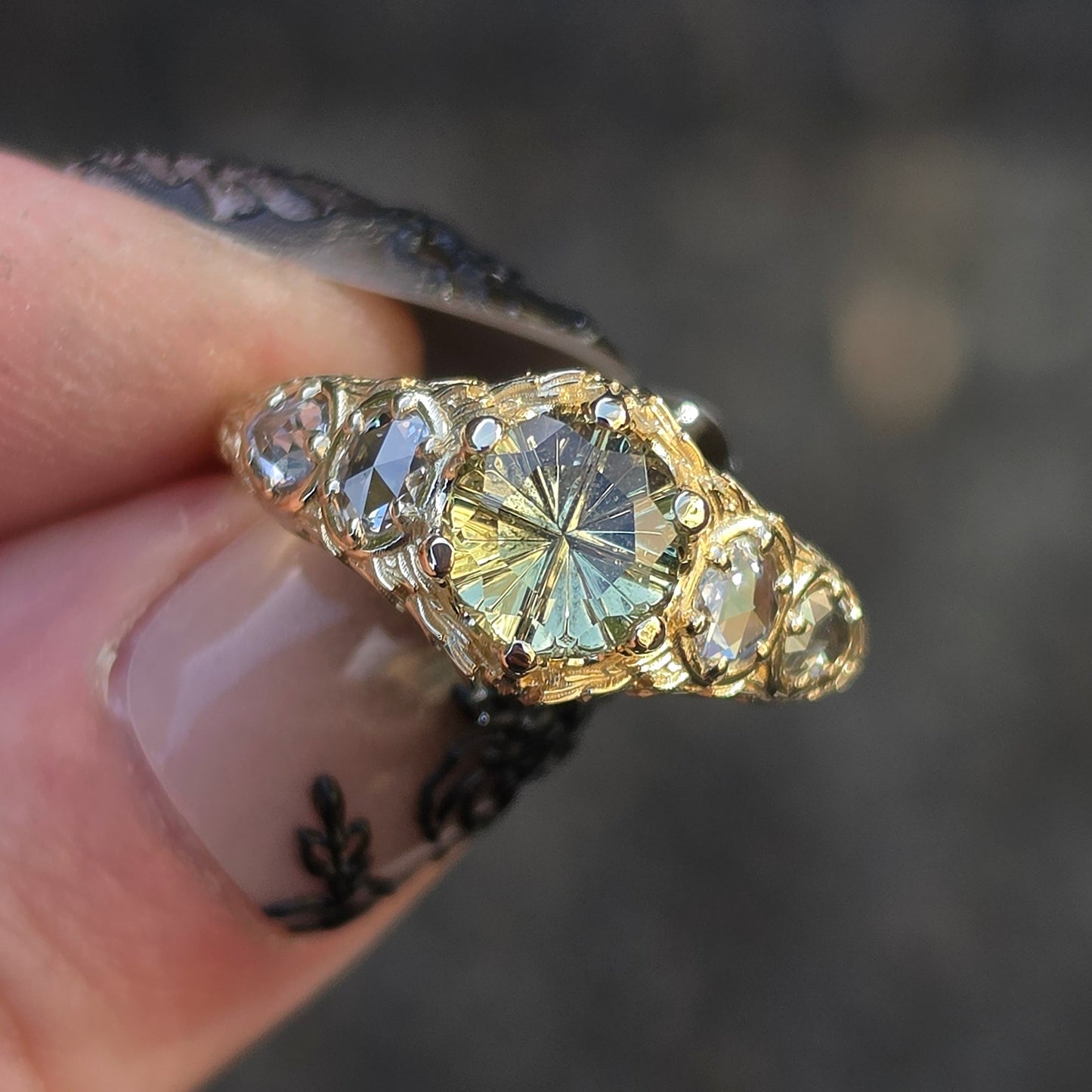A gold ring with a large yellow round center stone and smaller clear stones on either side, held between fingers against a blurred dark gray background.