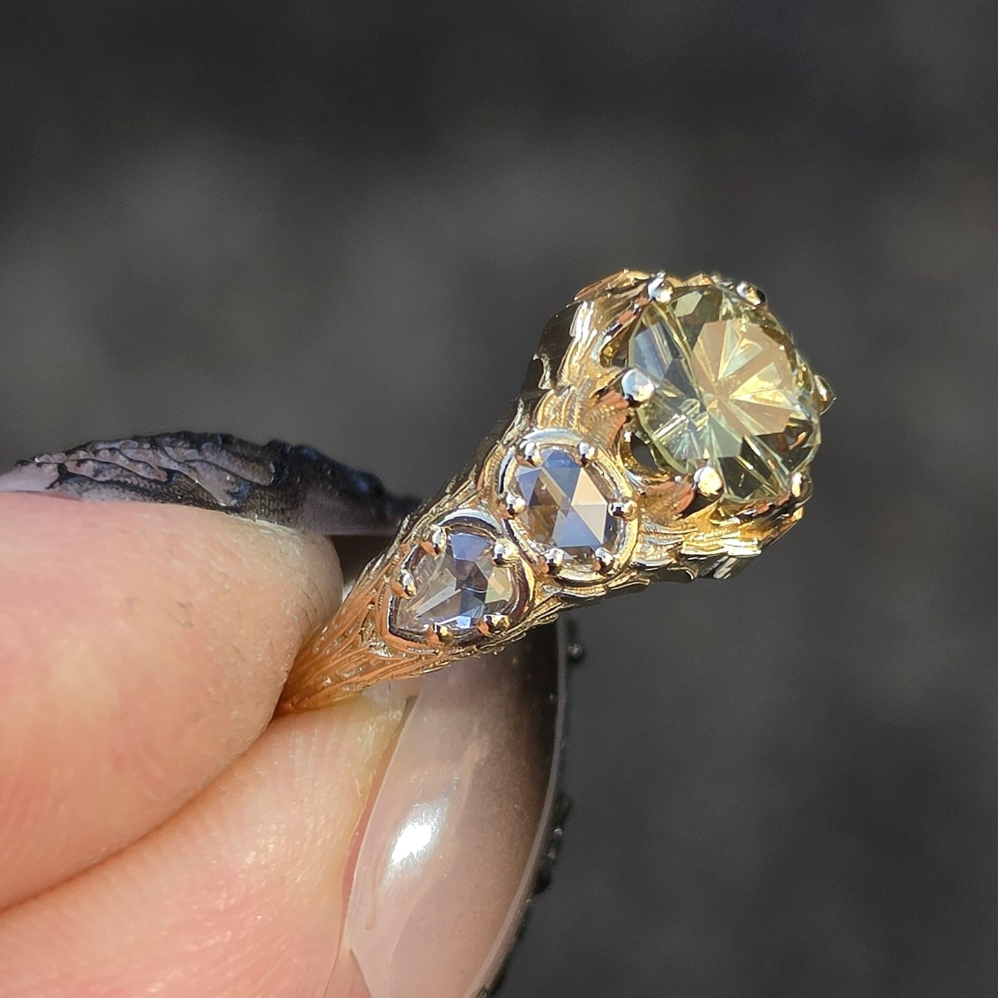 A close-up of a hand holding a gold ring with three clear gemstones, set against a dark, blurred background.