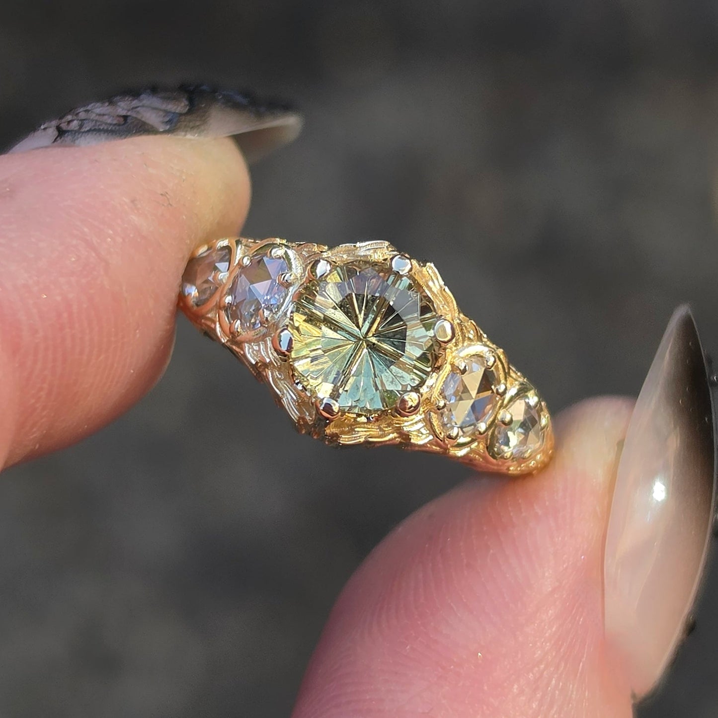 A gold ring with a large round iridescent green gemstone and smaller clear stones, held between fingers against a blurred dark gray background.