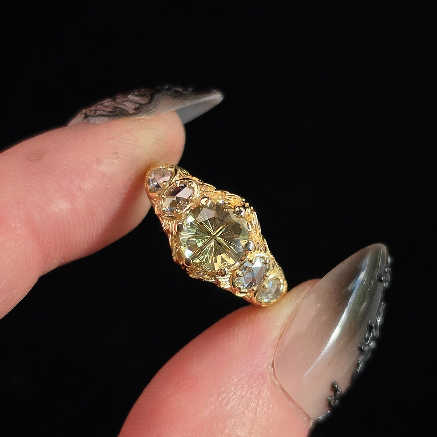 A close-up of a vintage-style gold ring with a large oval pale yellow center stone and smaller clear stones on either side, held between two fingers against a solid black background.
