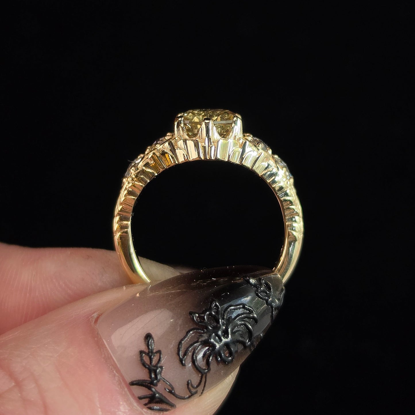 A close-up of a yellow gold ring with a textured, beaded band and a small round center stone, held between fingers against a solid black background.