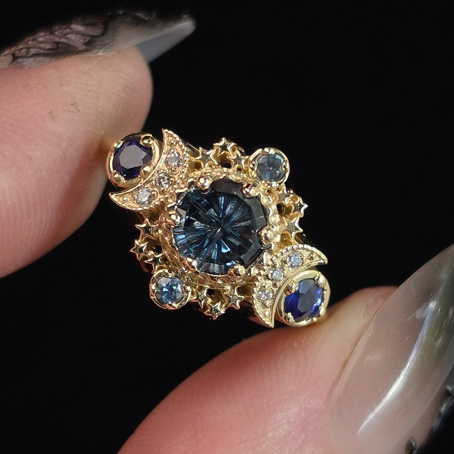 A close-up of a hand with long manicured nails holding an ornate gold ring featuring a central round dark blue gemstone and smaller blue gemstones, set against a solid black background.