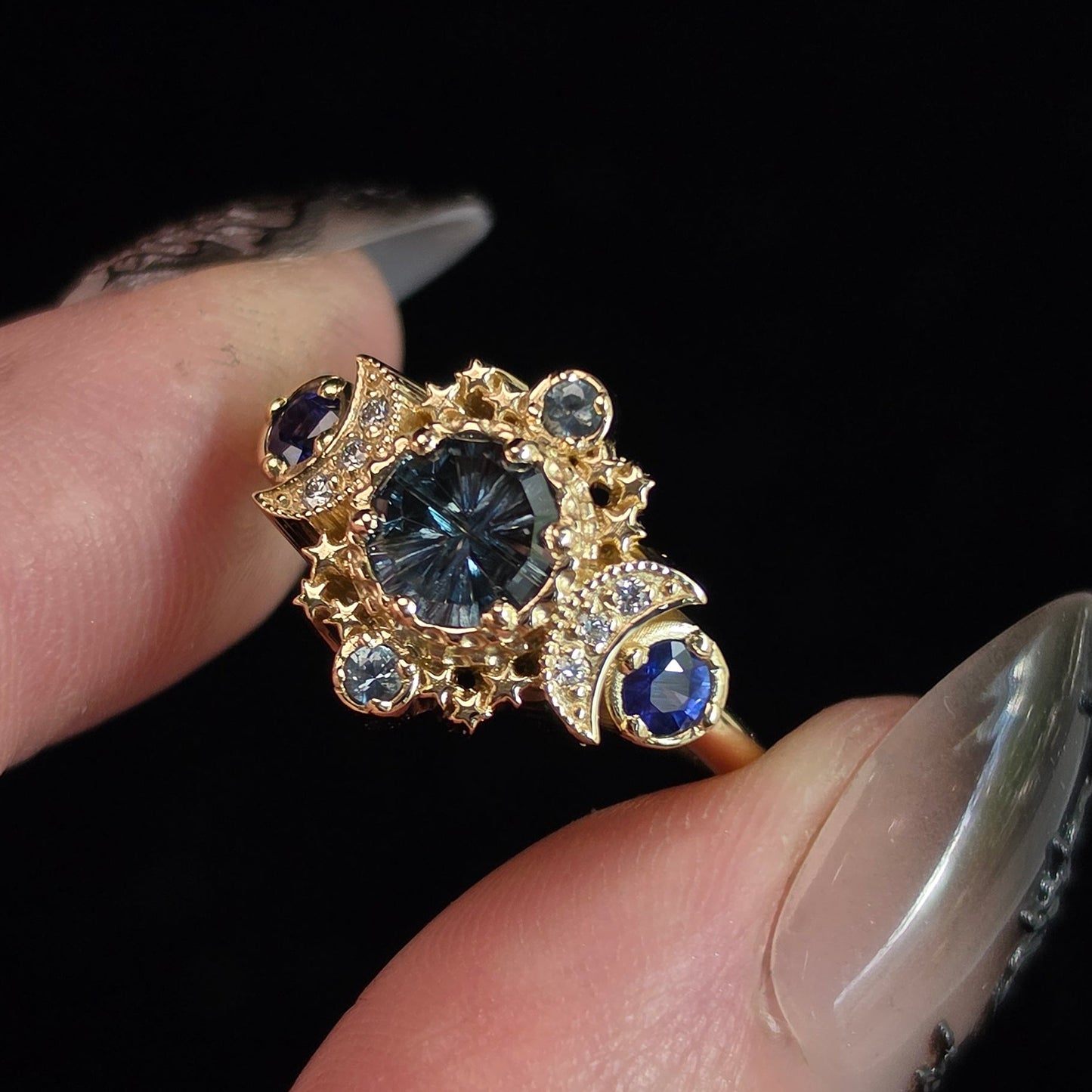 A close-up of a hand with long manicured nails holding a gold ring featuring a large round dark blue center stone and smaller blue and clear accent stones, set against a solid black background.