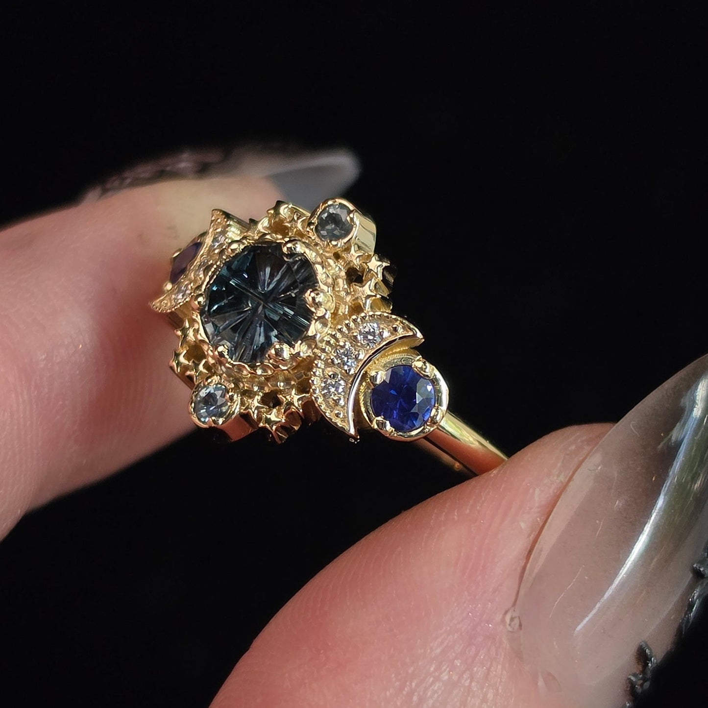 A close-up of a hand wearing a gold ring with a large round dark gemstone and smaller blue gemstones, set against a solid black background.