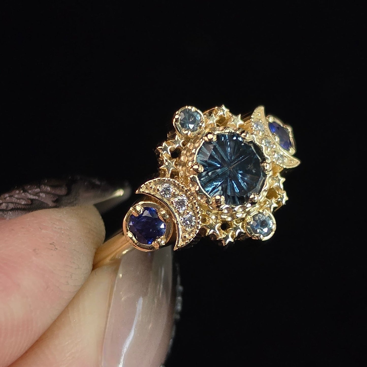 A gold ring with a central round blue gemstone and smaller blue and clear accent stones, held between fingers against a solid black background.