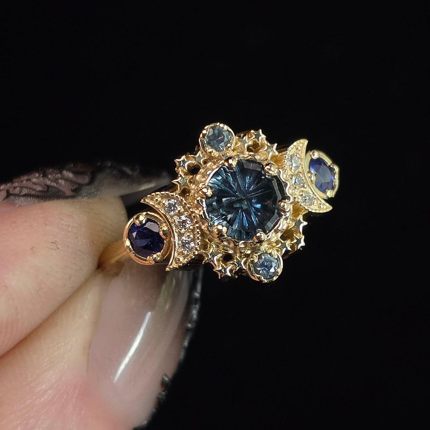 A gold ring with a large round blue gemstone and smaller blue and dark blue stones, held between fingers against a solid black background.