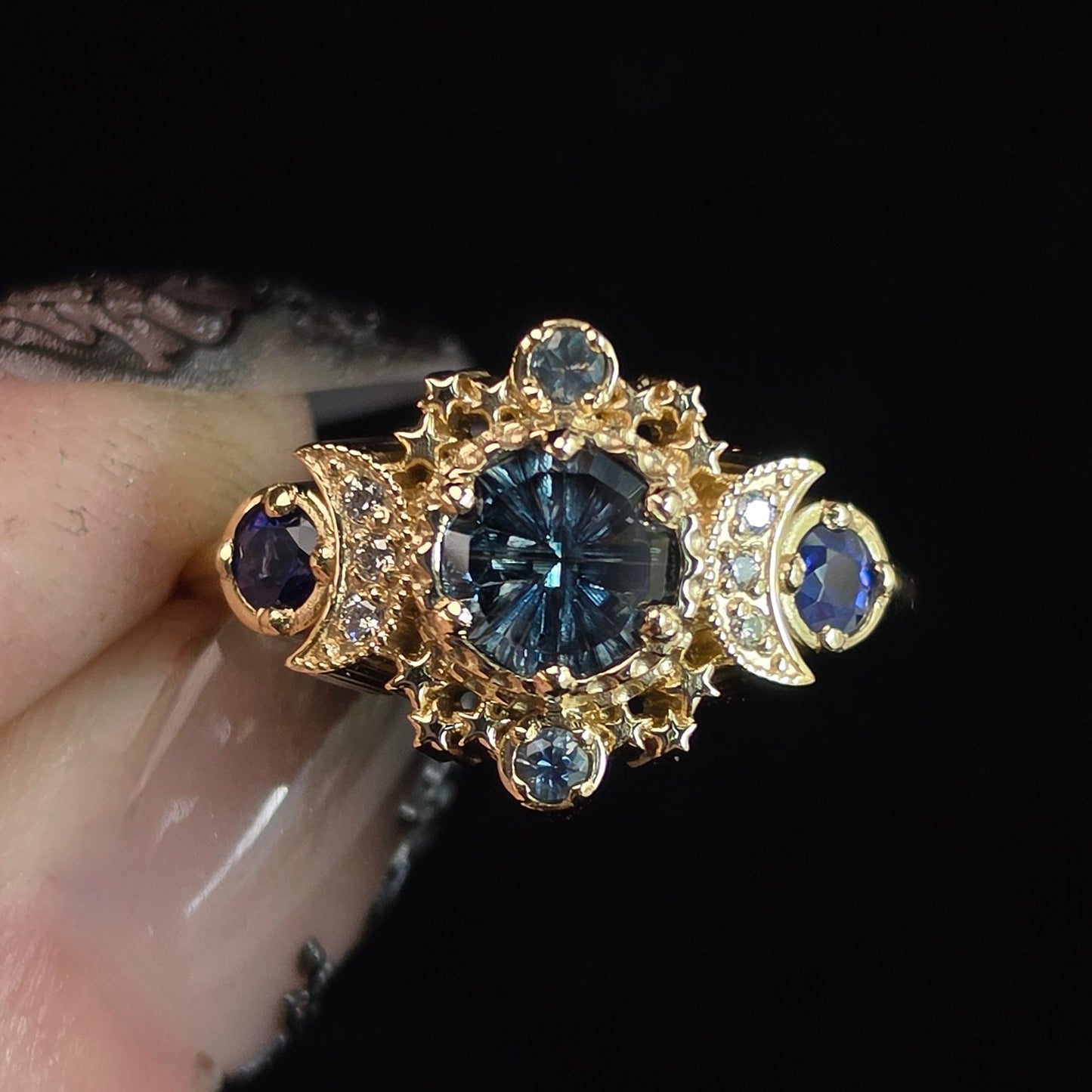 A gold ring with a large round dark blue center stone and smaller blue stones, held between fingers against a solid black background.