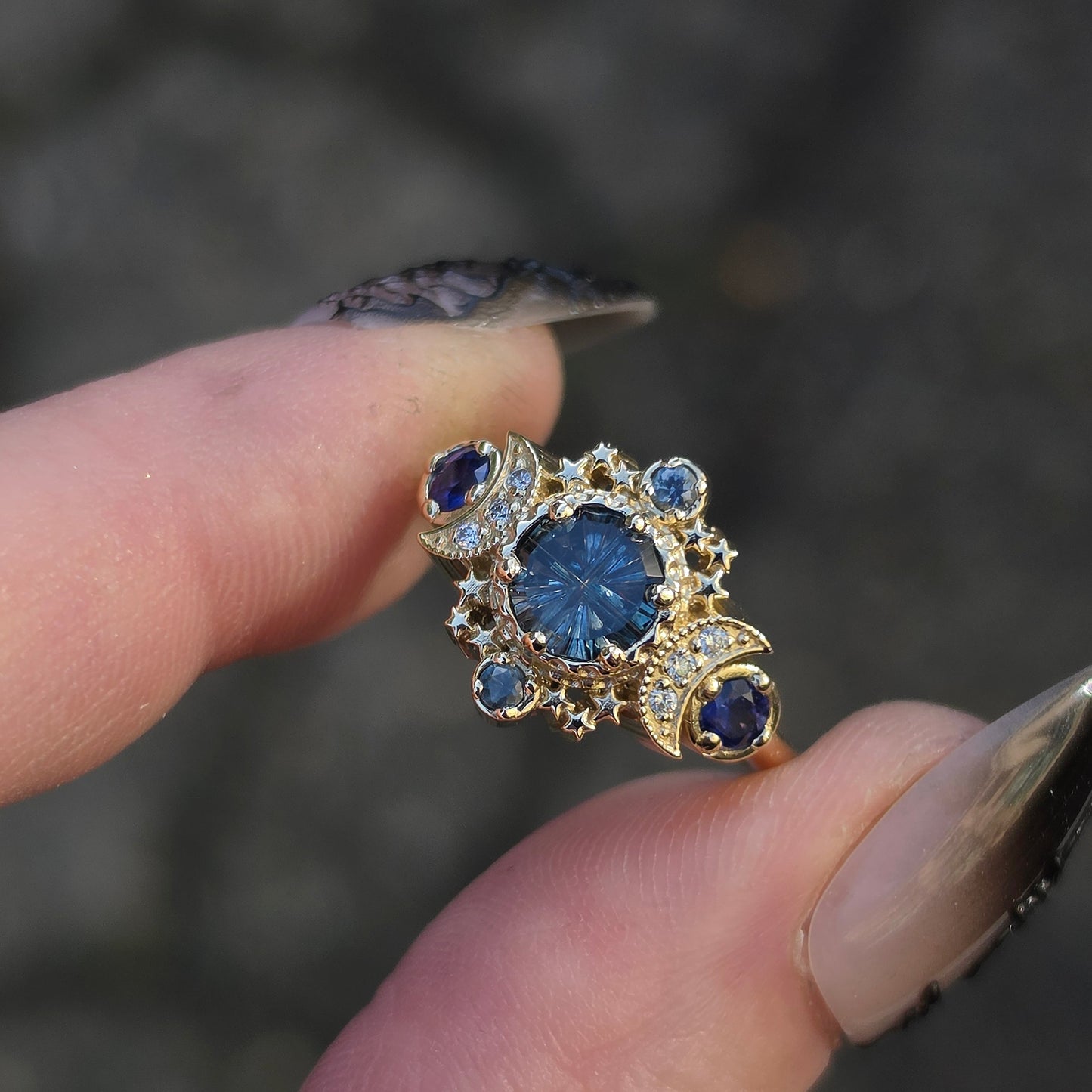 A close-up of a hand holding a gold ring with a large round blue center stone and smaller blue stones around it, against a blurred gray outdoor background.