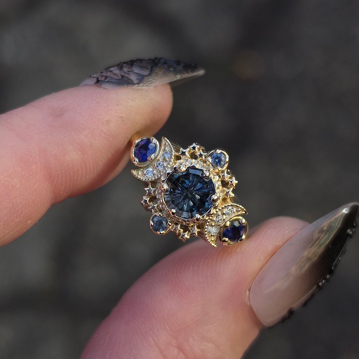 A close-up shot of a hand with long manicured nails holding a gold ring featuring a central round-cut dark blue gemstone surrounded by smaller blue and clear stones.