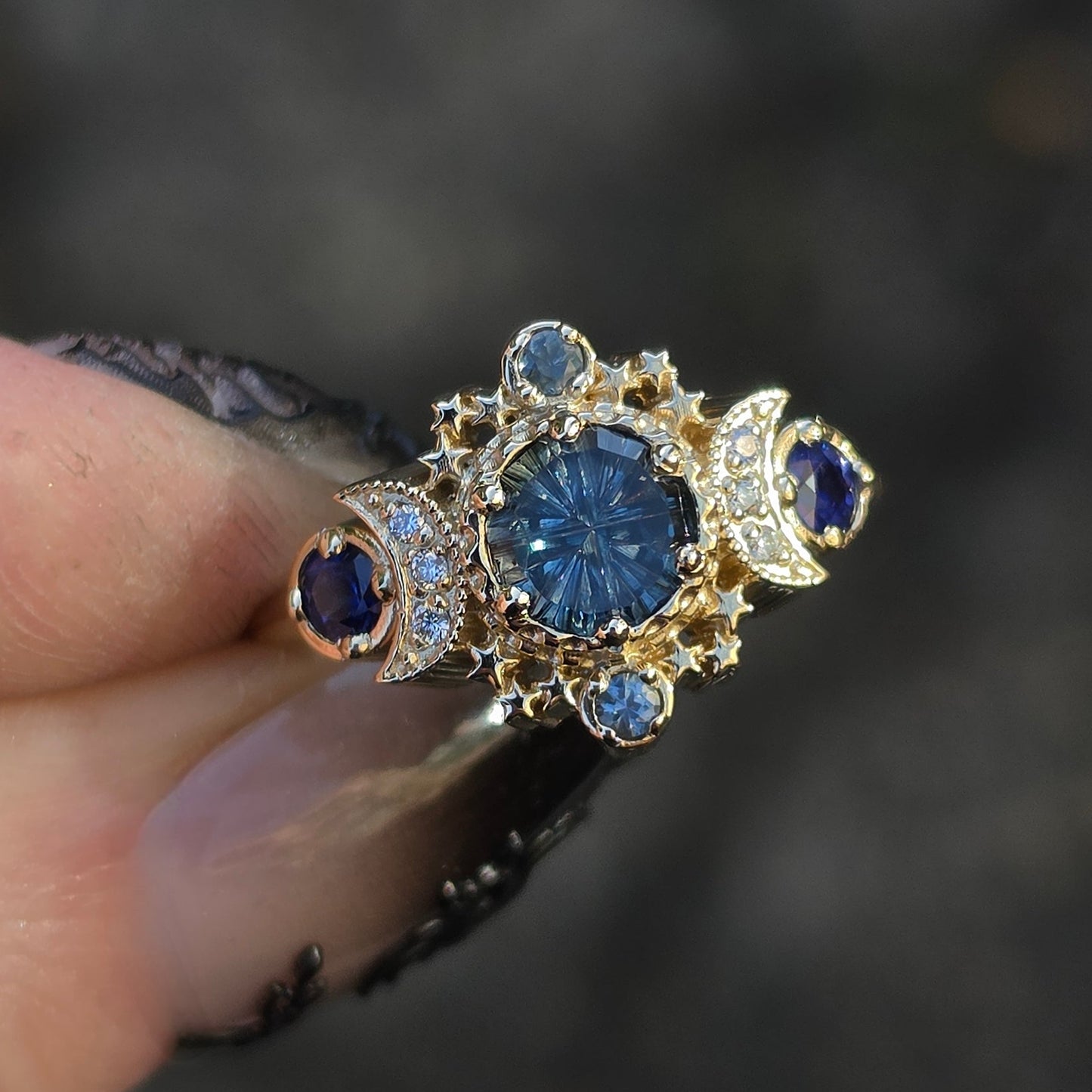 A close-up of a hand holding a vintage-style ring with a large round blue center stone, surrounded by gold-toned filigree and small blue accent stones, against a blurred dark gray background.